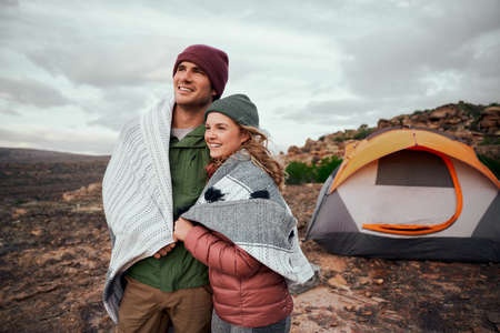 Romantic man and woman wrapped in a blanket standing together outside tent on mountain looking at...