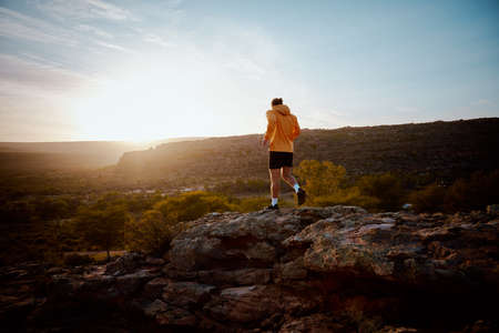 Rear view of young fitness male athlete running through rocky mountain pathの写真素材