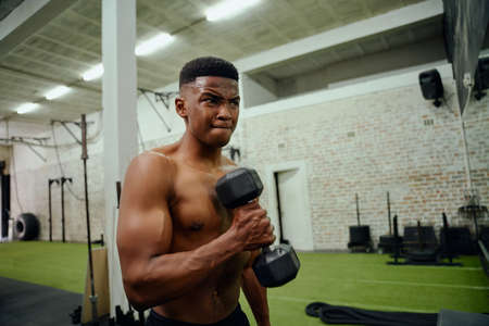 African American male holding weights during an intense workout. Mixed race male looking at himself in the gym mirror while lifting dumbbells. High quality photoの写真素材