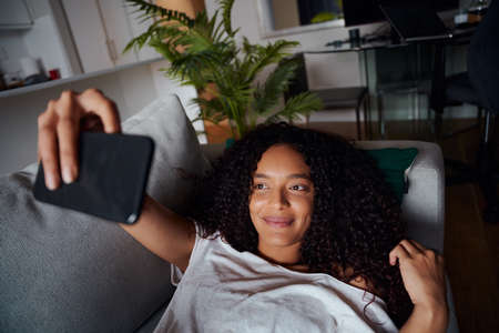 Mixed race female taking selfies on cellular device while lying on couch smiling. High quality photoの写真素材