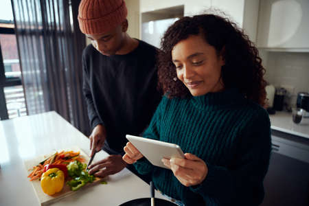 Happy, young mixed race couple cooking food in the kitchen and holding a tablet. Woman looking at tablet for food recipe. High quality photoの写真素材