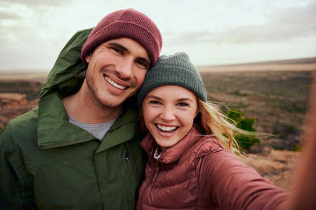 Portrait of cheerful young woman embracing man taking selfie during camping on mountainの写真素材
