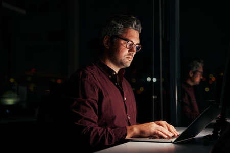 Serious businessman sitting near window working on laptop late at night in officeの写真素材