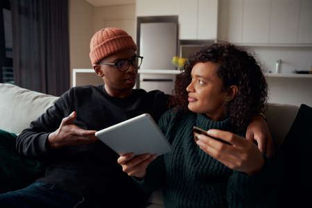Multi-ethnic couple fighting about purchasing goods online on tablet using credit card, sitting on sofaの写真素材