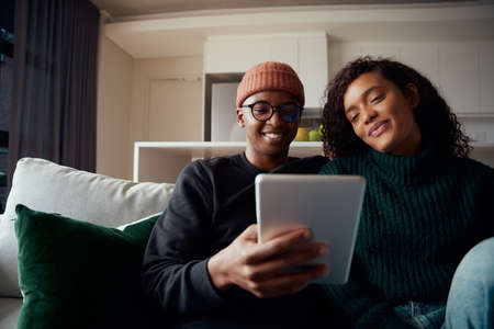 Young adult couple, multi-ethnic, sitting on sofa in apartment using tablet.の写真素材