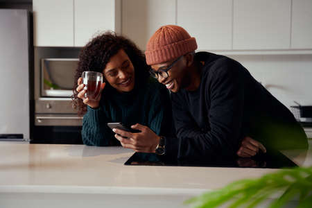 Biracial couple laughing at phone while in kitchen in modern apartment