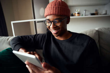 Happy adult black male smiling while looking at tablet. Modern apartment on the sofa.の写真素材