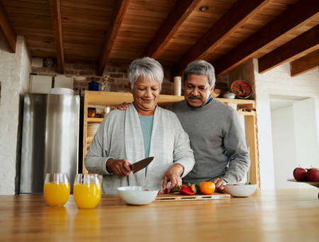 Multi-cultural elderly couple preparing fruit salad in modern kitchen. Healthy lifestyle at home. Orange juice and fruit.