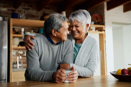 Happily retired elderly biracial couple smiling at each other. Wife holding husband in kitchen.