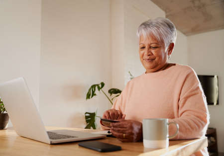 Elderly multi-ethnic female happily making online purchase on laptop, sitting at counter.