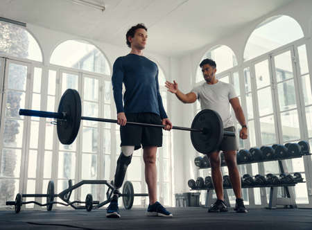 differently abled athlete weightlifting with his coach in the gym. Man with prosthetic leg being coached by his coachの写真素材