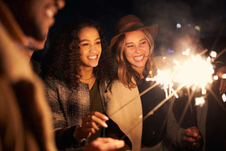 Multi-cultural best friends at a party smiling while lighting sparklers. Night time, stylish party.の写真素材