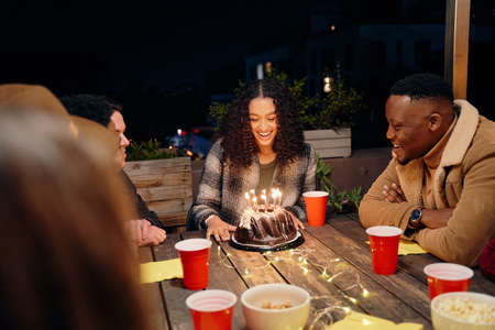Group of diverse young adults celebrating biracial female birthday, sitting at trendy rooftop party...