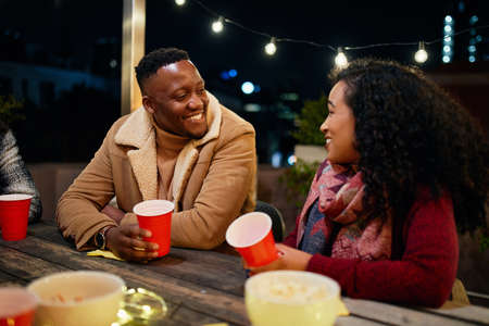 Biracial female and black male talking while at a rooftop party in the city. Trendy, drinking red...