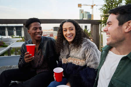 Group of multi-cultural young adults laughing at joke together while sitting at rooftop party in the...