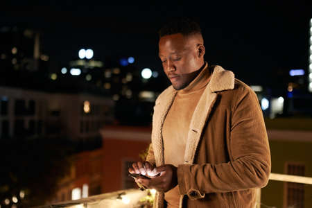 Independent, tall, handsome black male standing on rooftop terrace, texting on cellphone. Cityscape...