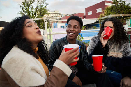 Group of multi-cultural young adults sitting on a rooftop terrace socializing, drinking and...