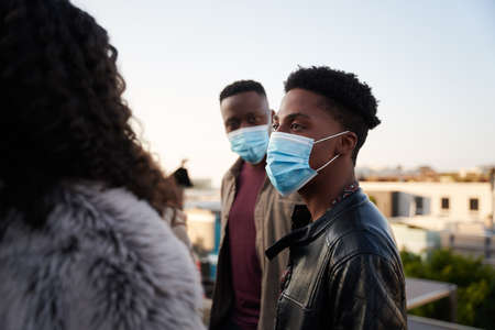 Black male and multi-culural friends chatting on rooftop terrace with masks on. City living and good times.の写真素材