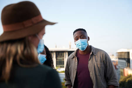 Black male and multi-cultural friends chatting on rooftop terrace with masks on. City living and...