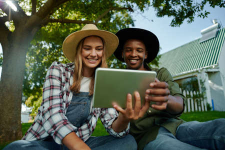 Mixed race male and female famers sitting together on green grass looking at digital tablet...