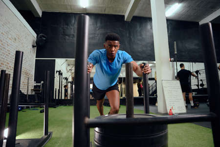 African American man working out intensely during cross fit training. Male athlete pushing the sled...