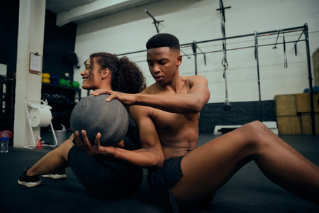 Multi-ethnic friends doing cross fit in the gym. African American male and female sitting down on the floor and passing a medicine ball during exercise routine. High quality photo. High quality photoの写真素材