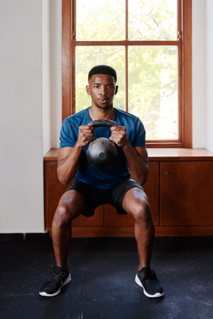 Determined young black man in sportswear doing kettlebell squats at the gymの写真素材