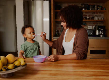 Biracial mother feeding daughter wearing casual clothing in kitchen at homeの写真素材