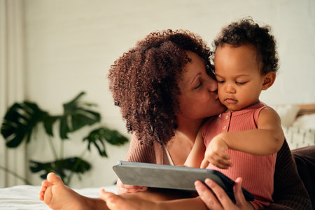 Happy biracial mother kissing daughter on cheek while using digital tablet on bed at homeの写真素材