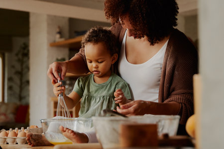 Biracial mother and daughter in casual clothing whisking eggs together in kitchen at homeの写真素材