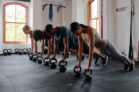 Group of determined multiracial young adults in sportswear doing plank exercise with kettlebell at the gymの写真素材