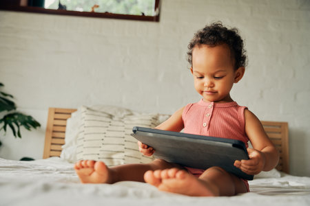 Focused biracial baby girl in casual clothing sitting on bed with digital tablet at homeの写真素材