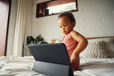 Biracial baby girl in casual clothing sitting on bed concentrating on digital tablet at homeの写真素材