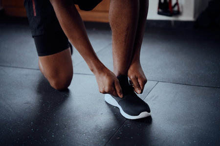 Close-up of young black man kneeling while tying shoelace of sports shoe at the gymの写真素材