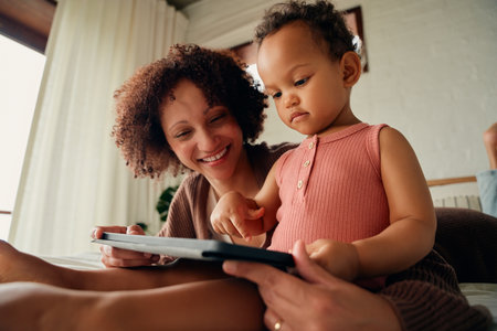 Biracial mother and daughter in casual clothing on bed playing with digital tablet at home
