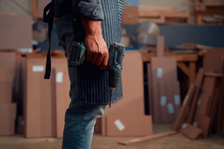 Close up of multiracial young man holding drill next to woodpile in woodworking factoryの写真素材