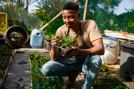 Happy young black man smiling while wearing apron and examining the plants at garden centerの写真素材