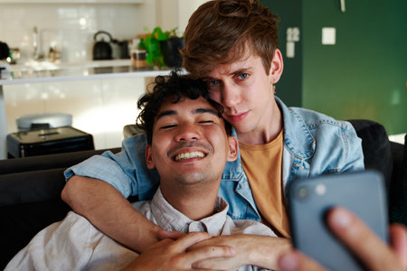 Young gay couple taking selfie with mobile phone while embracing on sofa in living room at homeの写真素材
