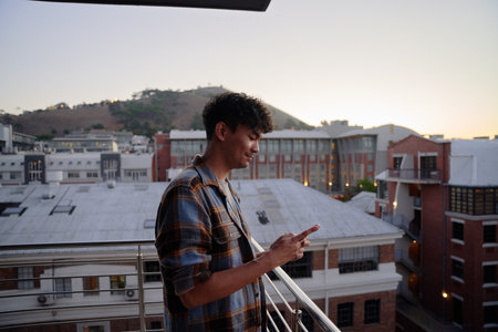 Profile view of young multiracial man in shirt using phone on balcony of apartment during sunsetの写真素材