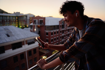 Profile view of young multiracial man in shirt smiling while using phone on balcony of apartment during sunsetの写真素材
