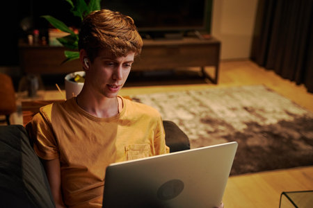 Young caucasian man wearing t-shirt while working on laptop with wireless headphones on sofa at homeの写真素材