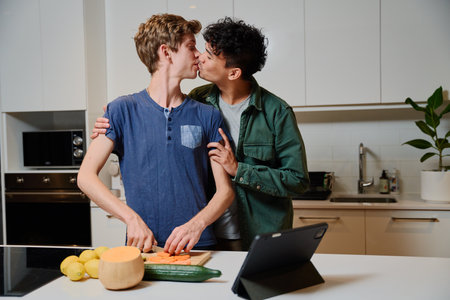 Young gay couple in casual clothing kissing while preparing dinner by digital tablet in kitchen at homeの写真素材