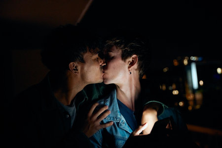 Young gay couple in casual clothing kissing while holding mobile phone on balcony of apartment at nightの写真素材