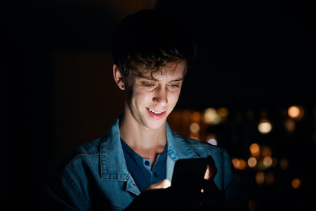 Young caucasian man in casual clothing smiling while texting on mobile phone outside apartment at nightの写真素材