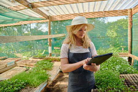 Young caucasian woman wearing apron and hat using digital tablet by plants in plant nursery