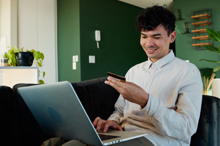 Young multiracial man in shirt smiling while using laptop and credit card on sofa in living room at homeの写真素材