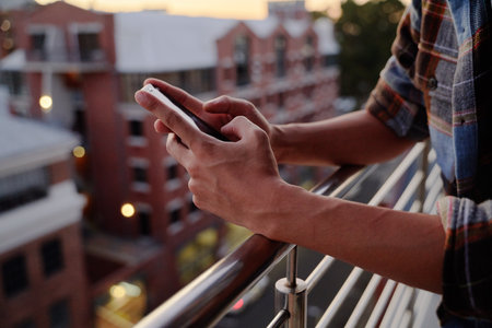 Close-up of young multiracial man in checkered shirt using phone on balcony of apartment during sunsetの写真素材