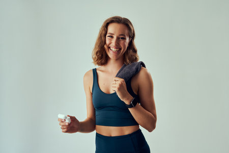 Happy young caucasian woman wearing sports clothing holding wireless headphones and towel in studioの写真素材
