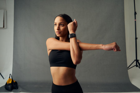 Focused young biracial woman wearing sports clothing and fitness tracker doing cross arm stretch in studioの写真素材