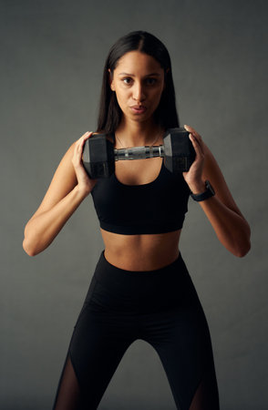 Focused young biracial woman wearing sports bra and leggings exhaling while holding weights in studioの写真素材